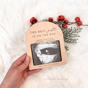May include: A wooden pregnancy announcement frame with the text "THE BEST gift IS ON THE WAY" and "MERRY CHRISTMAS GRANDMA + GRANDPA". The frame holds a sonogram image and reads "ARRIVING AUGUST 2026". Red bells and greenery decorate the background.
