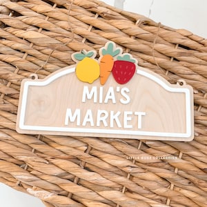May include: Wooden sign with the text "MIA'S MARKET" in white, featuring a lemon, carrot, and strawberry. The sign has a white border and is hanging by a twine string. The background is a woven basket.