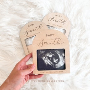 May include: Three wooden arch-shaped frames with a white background. The frames are engraved with the text "Baby Smith" and "Our Little Rainbow". The bottom frame has a black and white ultrasound image inside and the text "Arriving February 2025".