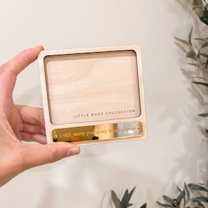 May include: A wooden photo frame from the Little Bugz Collection. The frame is rectangular with a light wood finish. A gold-coloured metal plate at the bottom has the inscription "I will carry you in my heart always."