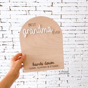 May include: Wooden sign with the text "Best grandma ever" in white letters and "hands down" in black letters. The sign also includes the text "Love, Elayna & Ethan" and "Little Bugz Collection".