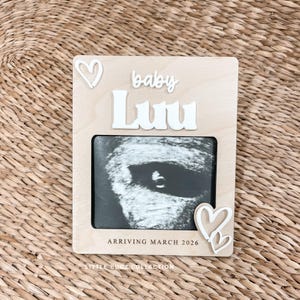May include: A wooden pregnancy announcement frame with the name "Luu" in white letters. The frame holds a sonogram image and is decorated with white hearts. The text "Arriving March 2026" is also visible.