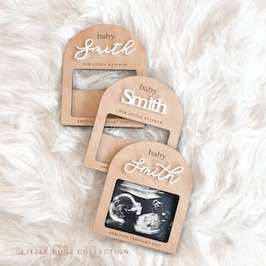 May include: Three wooden photo frames with a rounded arch top. Each frame has a white cutout of the word "baby" and a name, "Smith", with the text "Our Little Rainbow" underneath. The bottom frame has a black and white ultrasound image inside and the text "Arriving February 2025".