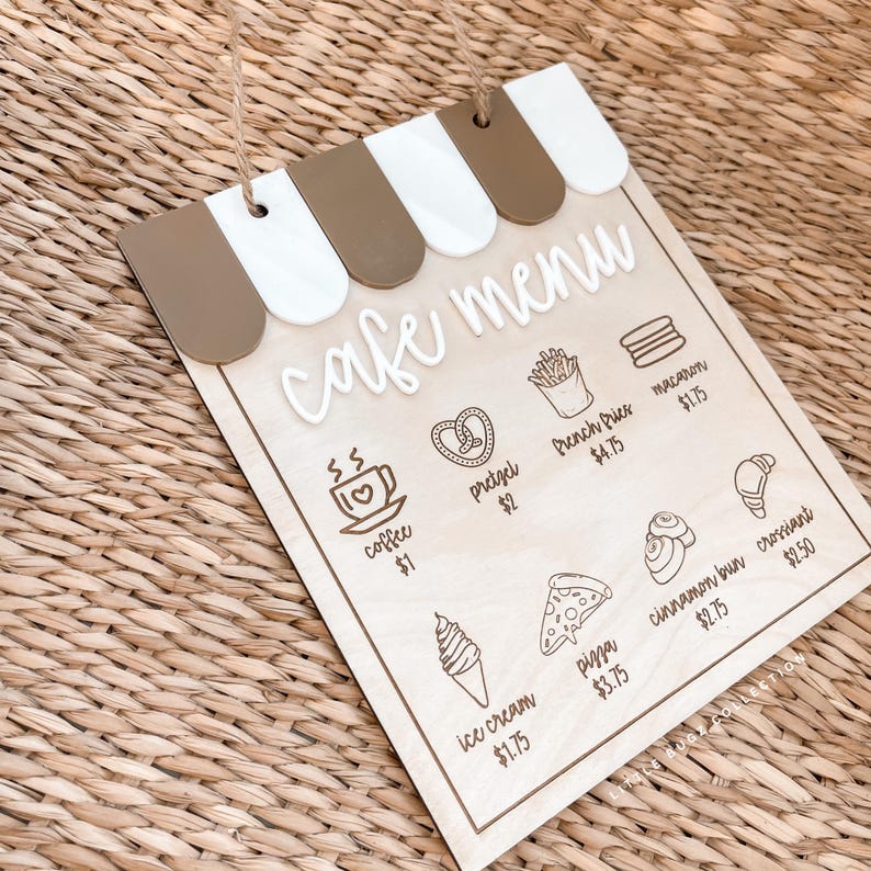 May include: Wooden cafe menu sign with a brown and white awning design. The sign features the words "cafe menu" in cursive and illustrations of coffee, pretzels, pizza, and other food items. The sign is hanging by a twine string.