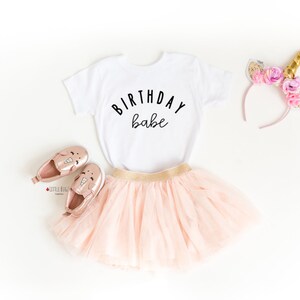 May include: A white t-shirt with the text "Birthday babe" in black lettering. The shirt is paired with a pink tulle skirt, gold shoes with unicorn designs, and a pink headband with gold flowers and a unicorn horn.