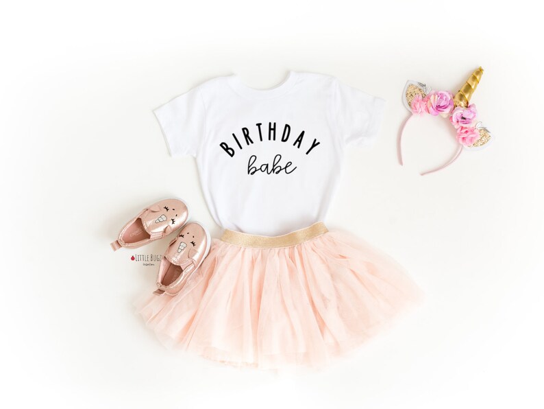 May include: A white t-shirt with the text "Birthday babe" in black lettering. The shirt is paired with a pink tulle skirt, gold shoes with unicorn designs, and a pink headband with gold flowers and a unicorn horn.