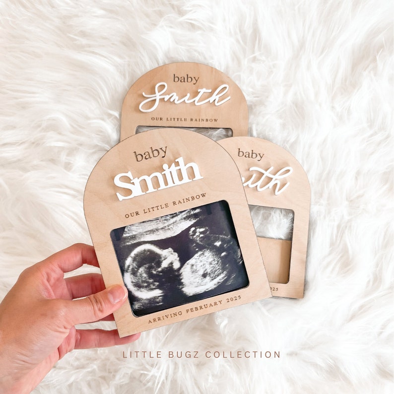 May include: A wooden sign with a white arch and the text "baby Smith OUR LITTLE RAINBOW" and a black and white ultrasound image. The sign also says "ARRIVING FEBRUARY 2025".