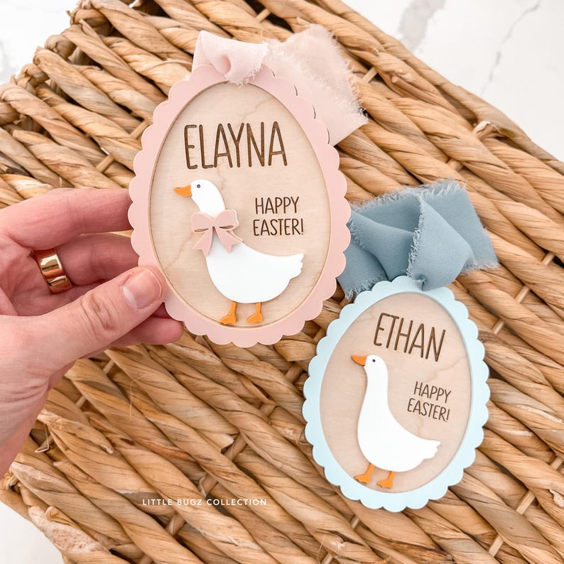 Puede incluir: Dos adornos de madera con forma de huevo de Pascua con bordes festoneados. Uno es rosa y el otro azul. Cada uno presenta un ganso blanco con pico y patas naranjas, un lazo y el texto "Elayna" y "Ethan" con "&iexcl;Felices Pascuas!"