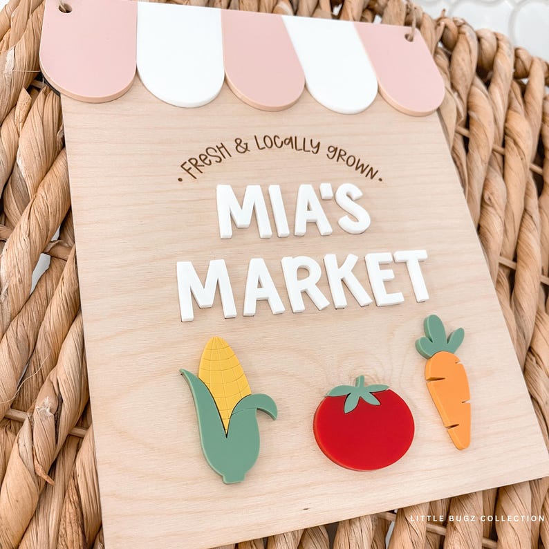 Puede incluir: Letrero de madera con un dise&ntilde;o de toldo rosa y blanco. El letrero dice "Fresh & Locally Grown. MIA'S MARKET" en letras blancas. Incluye recortes de verduras coloridas de ma&iacute;z, un tomate y una zanahoria.