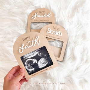May include: A wooden photo frame with a white arch and the text "baby Smith OUR LITTLE RAINBOW" in a cursive font. The frame has a space for a photo and the text "ARRIVING FEBRUARY 2025" is printed below the photo space. The frame is on a white fuzzy surface.