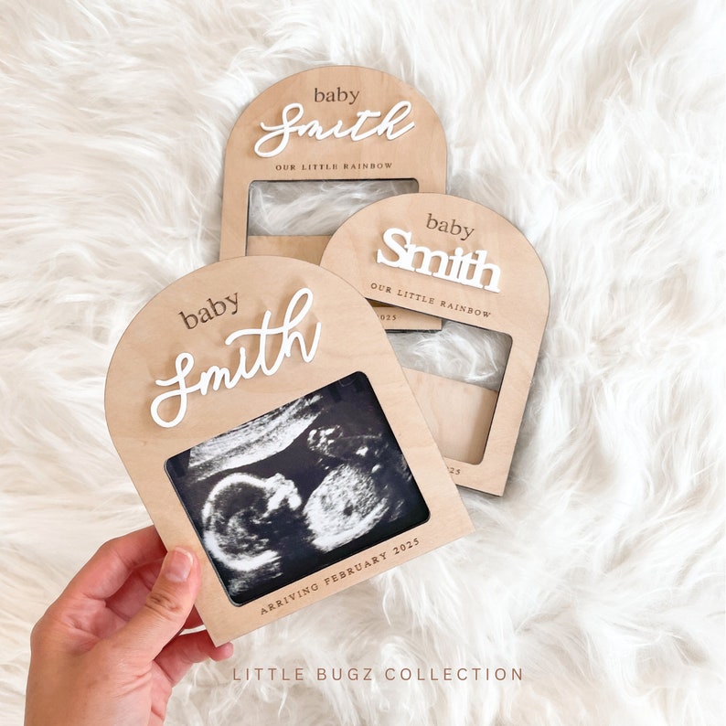 May include: A wooden photo frame with a white arch and the text "baby Smith OUR LITTLE RAINBOW" in a cursive font. The frame has a space for a photo and the text "ARRIVING FEBRUARY 2025" is printed below the photo space. The frame is on a white fuzzy surface.