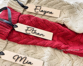 May include: Close-up of three personalized wooden gift tags with black lettering: "Elayna", "Ethan", and "Mia". The tags are attached to a beige and red cable-knit fabric with black ribbons. The words "Little Bugz Collection" are in the bottom right corner.