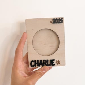 May include: A light-coloured wooden picture frame with a circular opening. The name "CHARLIE" is displayed in black letters at the bottom, with the year "2025" at the top. A paw print is next to the name. The frame is held by a hand.