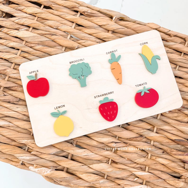 Puede incluir: Rompecabezas de madera con formas de frutas y verduras coloridas: manzana, br&oacute;coli, zanahoria, ma&iacute;z, tomate, fresa y lim&oacute;n. Cada forma est&aacute; etiquetada con su nombre. El texto "LITTLE BUG COLLECTION" est&aacute; en la parte inferior del tablero.