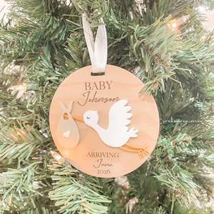 May include: A circular wooden Christmas decoration featuring a stork carrying a baby bundle. The ornament reads "BABY Johnson ARRIVING June 2026". A white ribbon is attached for hanging. The ornament is displayed on a Christmas tree.
