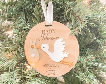 May include: A round wooden Christmas ornament with a stork carrying a baby bundle. The ornament has the text "BABY Johnson ARRIVING June 2026". A white ribbon is attached for hanging. The ornament is displayed on a Christmas tree.