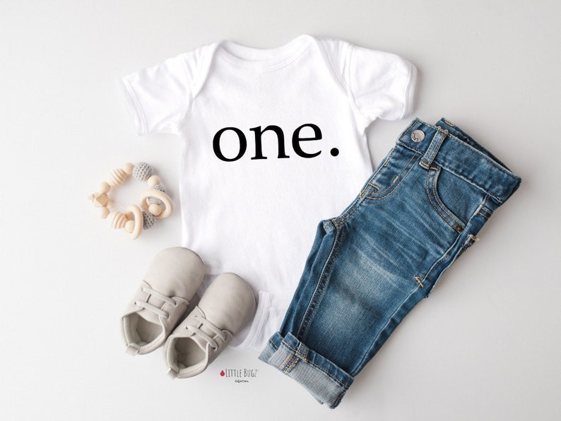 K&ouml;nnte beinhalten: Ein wei&szlig;er Baby-Strampler mit dem Wort "one" in Schwarz bedruckt. Der Strampler wird mit einer blauen Jeans und grauen Babyschuhen kombiniert.