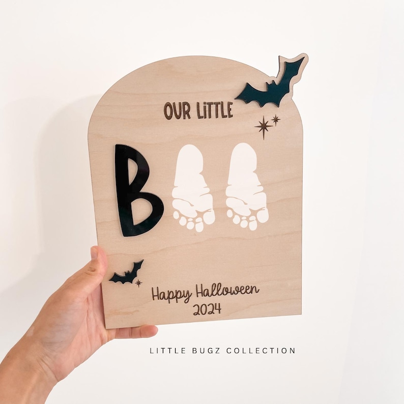 May include: A wooden sign with the text "Our Little Boo" and two white baby footprints. The sign also includes the text "Happy Halloween 2024" and a black bat cutout.