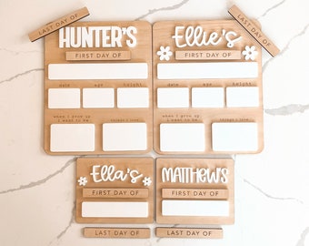 May include: Four wooden signs with white lettering, each with a child's name and the words "First Day Of" and "Last Day Of". The signs have spaces for writing the date, age, height, what the child wants to be when they grow up, and things they love.