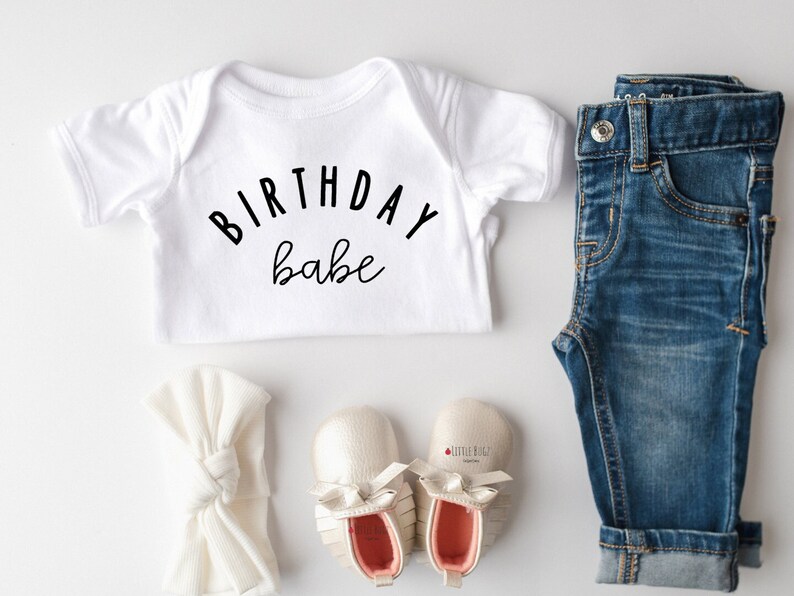 May include: White baby onesie with the text "Birthday babe" in black lettering. The onesie is paired with a white headband, blue jeans, and a pair of beige baby shoes.