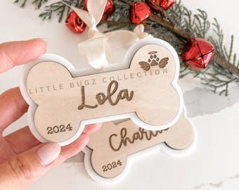 May include: Two wooden dog bone ornaments with white outlines. The top ornament has the text "Little Bugz Collection" and "Lola" with the year "2024" below. The bottom ornament has the text "Charlie" with the year "2024" below.
