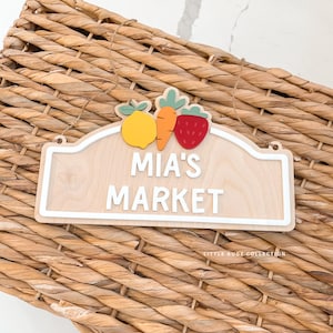 May include: Wooden sign with the text "MIA'S MARKET" in white letters. The sign is decorated with a lemon, carrot, and strawberry. The sign is hanging on a woven basket.