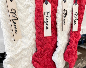 May include: Four cable-knit Christmas stockings, two red and two white, hang with wooden name tags. The tags read "Mama", "Elayna", "Ethan", and "Mia". The stockings are a festive holiday decoration.