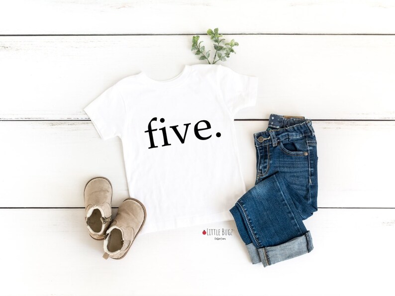 K&ouml;nnte beinhalten: Wei&szlig;es T-Shirt mit dem Wort "five" in Schwarz auf wei&szlig;em Holzgrund gedruckt. Das Shirt wird mit einer blauen Jeans und braunen Stiefeln kombiniert.