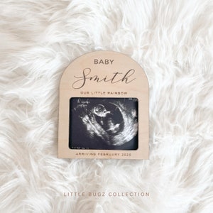 May include: Wooden frame with an arched top, featuring the text "Baby Smith Our Little Rainbow" and a black and white ultrasound image. The frame also includes the text "Arriving February 2025" and the words "Little Bugz Collection".