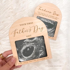 Op de afbeelding: Twee houten Vaderdagcadeaus met echografiebeelden. De frames hebben gewelfde toppen en bevatten de tekst "YOUR FIRST Father's Day GIFT" en "BABY SMITH ARRIVING DECEMBER 2026". Eén frame luidt "THIS YEAR WILL BE IN MOMMY'S BELLY NEXT YEAR I WILL BE IN YOUR ARMS".