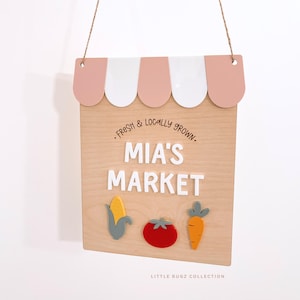 Puede incluir: Letrero de madera para "Mia's Market" con un dise&ntilde;o de techo festoneado en rosa y blanco. El letrero presenta el texto "Fresh & Locally Grown" y el nombre "MIA'S MARKET" en letras blancas. Debajo hay recortes de madera de ma&iacute;z, tomate y zanahoria.