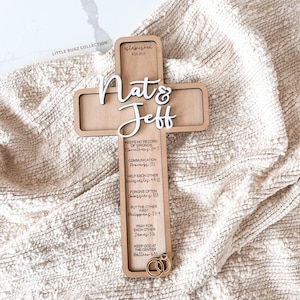 Peut inclure: Une croix murale en bois clair avec les prénoms Nat & Jeff en écriture blanche, le texte established 8.25.2015, et six versets bibliques dont 1 Corinthians 13:4-7, Proverbs 15:1, Ecclesiastes 4:9-10, Colossians 3:13, Philippians 2:3-4, James 5:16 et Matthew 6:33, avec deux anneaux dorés à la base.
