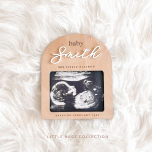 May include: A wooden arch-shaped frame with a white background and the text "baby Smith our little rainbow arriving February 2025" and a black and white ultrasound image inside. The frame is on a white fluffy surface.
