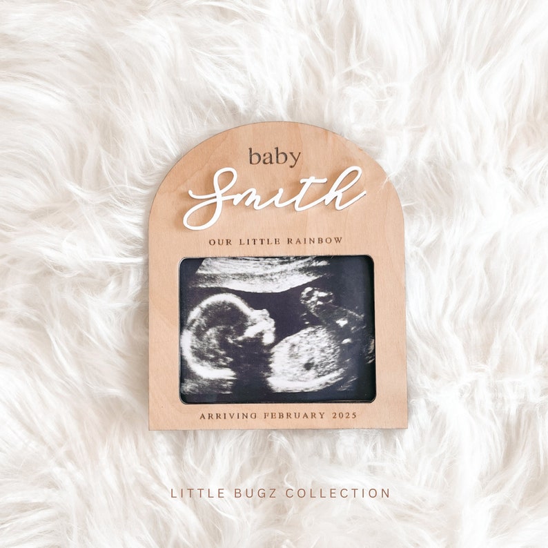 May include: A wooden arch-shaped frame with a white background and the text "baby Smith our little rainbow arriving February 2025" and a black and white ultrasound image inside. The frame is on a white fluffy surface.