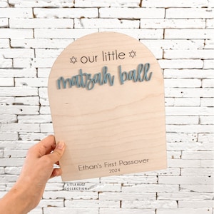 May include: A wooden sign with a blue arch and the text "our little matzah ball" with two stars. The sign also has the text "Ethan's First Passover 2024" and "Little Bugz Collection".
