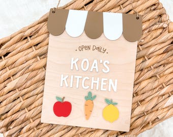 Puede incluir: Letrero de madera con diseño de toldo marrón y blanco, el texto "OPEN DAILY" y "KOA'S KITCHEN" en letras blancas. Presenta una manzana roja, una zanahoria naranja y un limón amarillo. Incluye una cuerda de yute para colgar.