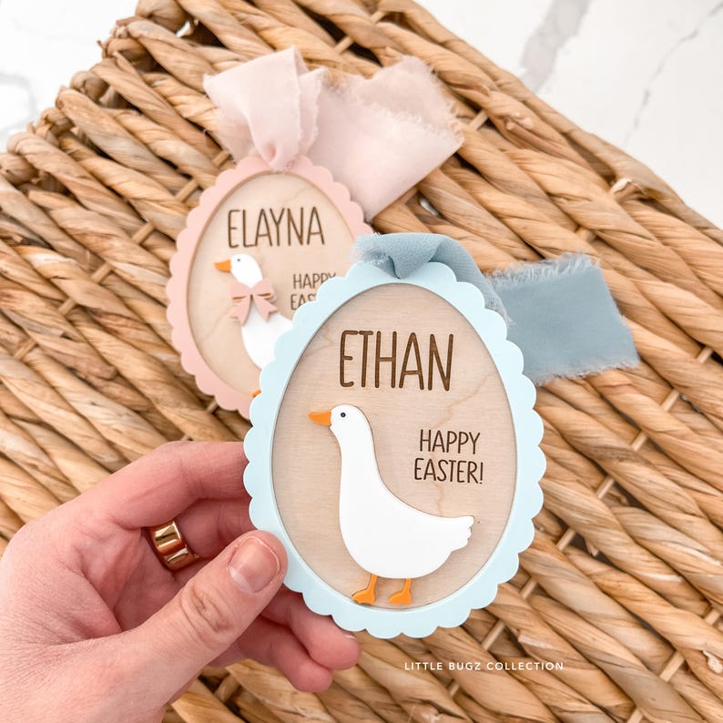 Puede incluir: Dos adornos de madera con forma de huevo de Pascua con bordes festoneados. Uno es rosa y el otro azul. Cada uno presenta un ganso blanco, un nombre y el texto "Happy Easter!". Se adjuntan cintas para colgar. El texto "Little Bugz Collection" est&aacute; en la parte inferior.
