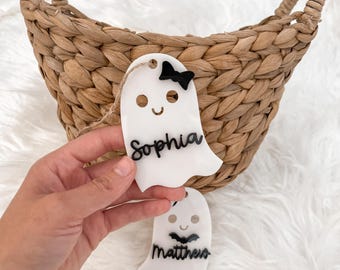 Op de afbeelding: Witte spookvormige naamlabels met zwarte tekst, een zwarte strik en een lachend gezicht. Op één label staat "Sophia", op de andere "Matthew". De labels worden voor een gevlochten mand en een pluizig wit oppervlak gehouden.