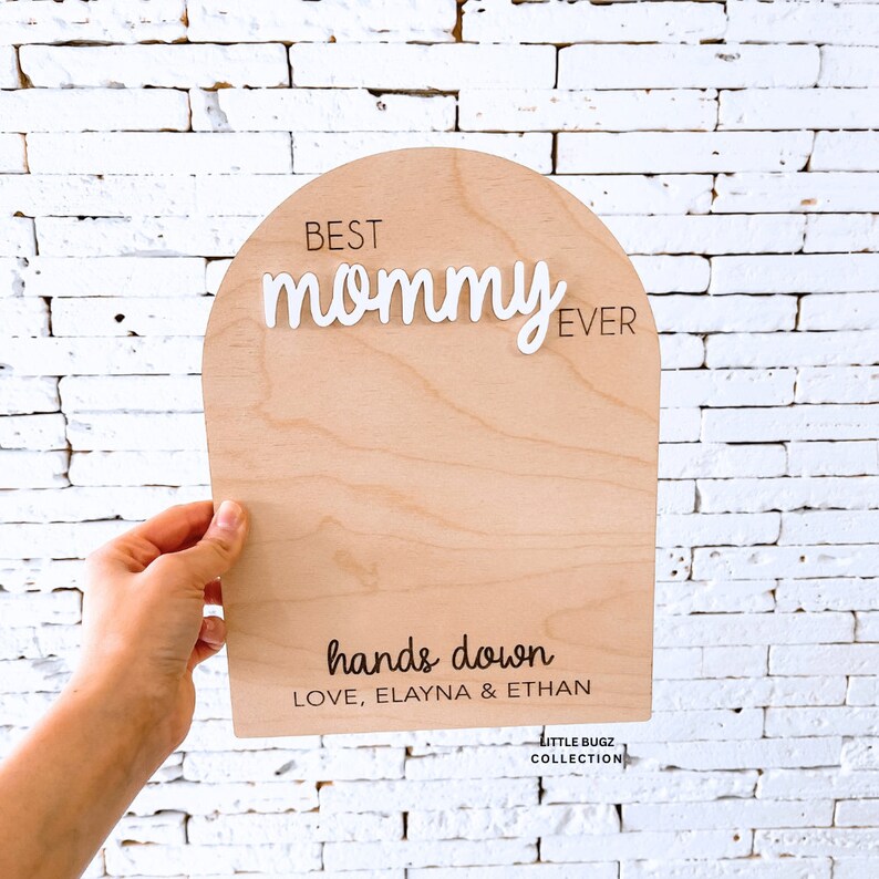 May include: Wooden sign with an arch shape, painted in a light wood color. The sign says "Best Mommy Ever" in white letters and "hands down" in black letters. Below the text, it says "Love, Elayna & Ethan".