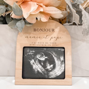 May include: A wooden frame with the words "BONJOUR mamie et papi" and "J'AI HÂTE DE VOUS RENCONTRER EN 2026!" is displayed. The frame holds a sonogram image. The frame is decorated with flowers.