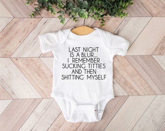 May include: White baby bodysuit with the text "LAST NIGHT IS A BLUR... I REMEMBER SUCKING TITTIES AND THEN SHITTING MYSELF" in black. The bodysuit is suspended on a wooden hanger against a light brown and white wooden backdrop.