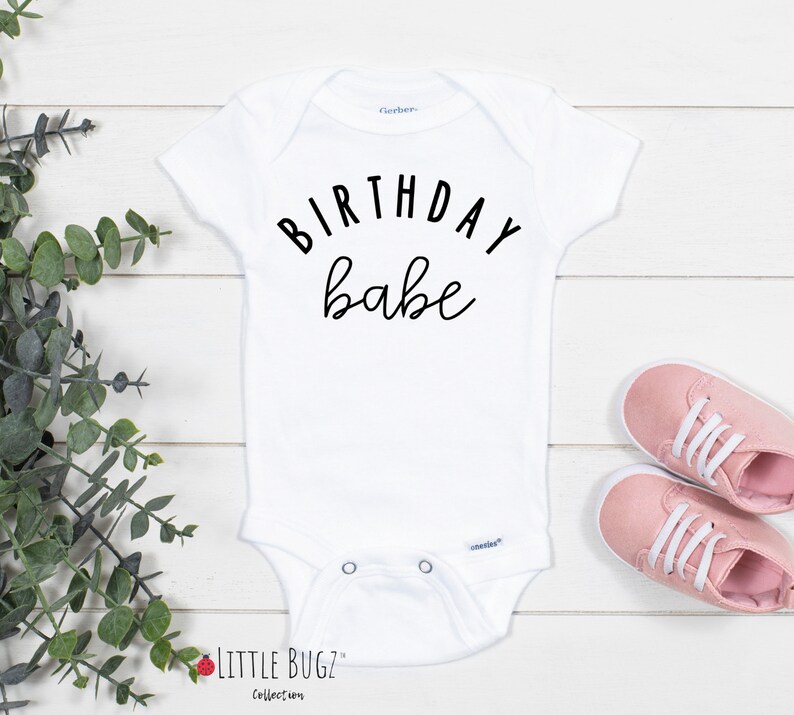 May include: White baby onesie with black text that reads "Birthday babe".