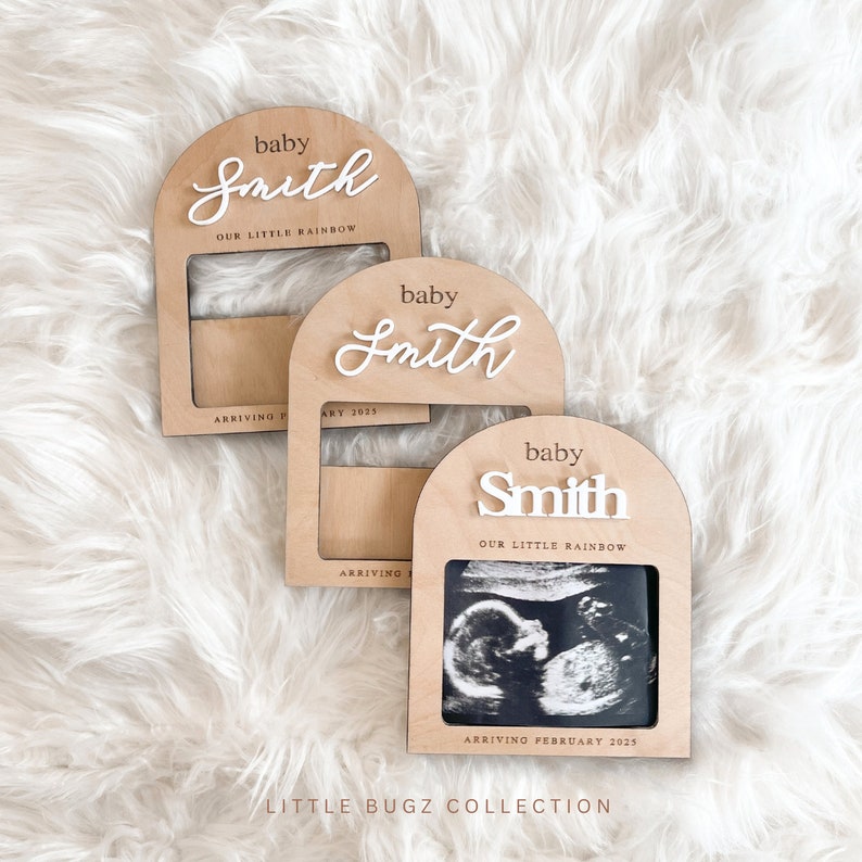May include: Three wooden arch-shaped photo frames with a cutout for a photo. The frames are light brown and have the text "baby Smith" and "our little rainbow" on them. The frames are on a white fluffy surface.