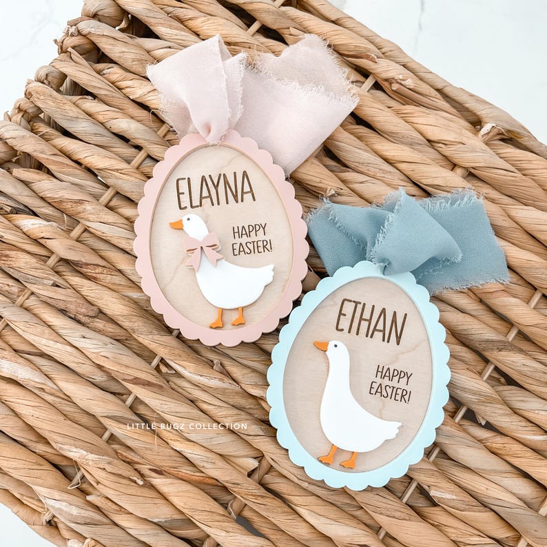 Puede incluir: Dos adornos de madera con forma de huevo de Pascua con bordes festoneados, cada uno con una ilustraci&oacute;n de ganso blanco y las palabras "Happy Easter!". Uno es rosa con una cinta rosa, el otro es azul con una cinta azul.