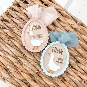 Puede incluir: Dos adornos de madera con forma de huevo de Pascua con bordes festoneados. Uno es rosa y el otro azul. Cada uno presenta un ganso blanco con patas naranjas y un lazo, un nombre y el texto "&iexcl;Felices Pascuas!"