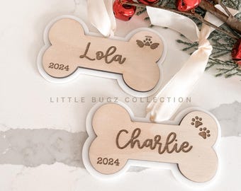 May include: Two bone-shaped wooden ornaments with white borders. One says "Lola" and the other says "Charlie" in cursive, with "2024" below. Paw prints and an angel wing design are also present. The ornaments are for pets.