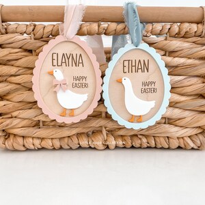 Puede incluir: Dos adornos de madera con forma de huevo de Pascua con bordes festoneados cuelgan de cintas. Uno es rosa con "ELAYNA" y un ganso blanco con un lazo. El otro es azul con "ETHAN" y un ganso. Ambos dicen "&iexcl;FELICES PASCUAS!"