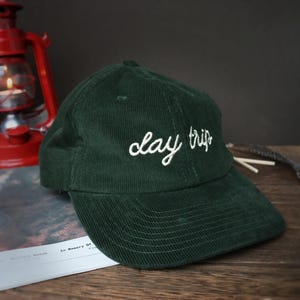 May include: A dark green corduroy baseball cap with white embroidery that reads "day trip".