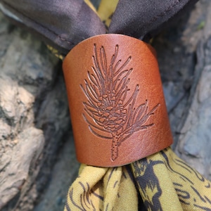 May include: A brown leather scarf ring featuring an embossed pine cone design. The ring encircles a scarf with a yellow and dark grey pattern. The background is a blurred rock surface.