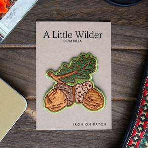 May include: An iron-on patch featuring an acorn with a leaf, in brown and green colours. The patch says "A Little Wilder Cumbria" and "Iron On Patch".